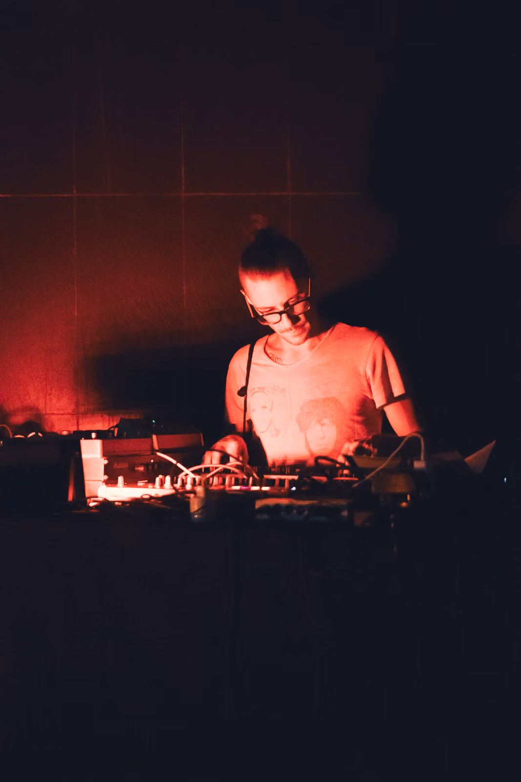 Una musicista con gli occhiali, illuminata di rosso, è china su un tavolo di sintetizzatori e mixer.