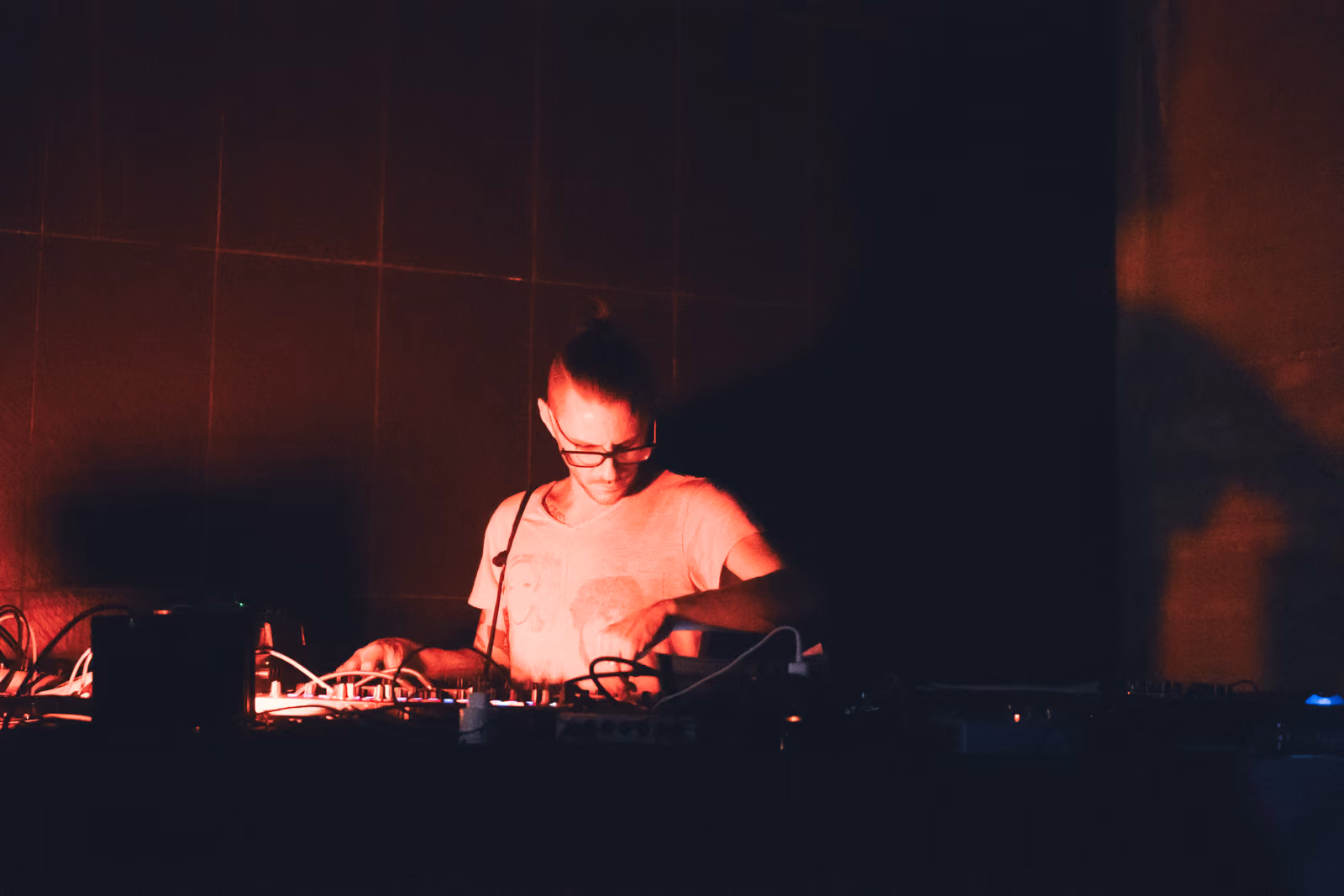 Un musicista con la barba e gli occhiali, illuminato di rosso, regola i comandi di un mixer.