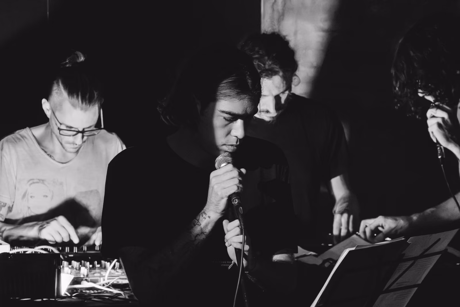 In bianco e nero, un cantante al microfono è affiancato da altri musicisti al mixer e alle tastiere.