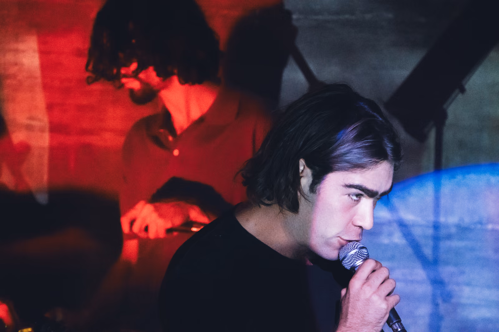 Un cantante con i capelli lunghi si china sul microfono, con un altro musicista sullo sfondo in una luce rossa.