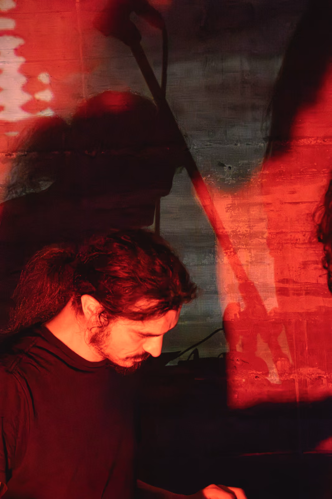 Un musicista con i capelli lunghi e mossi guarda verso il basso, illuminato da un fascio di luce rossa.