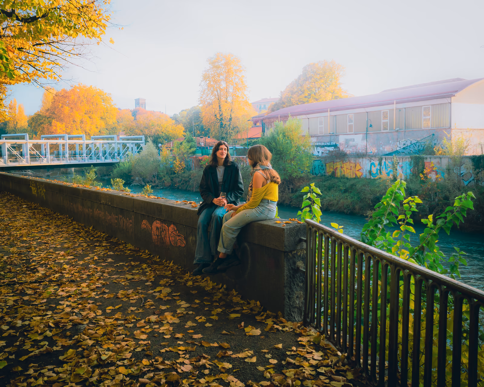 Due persone sedute su un muretto lungo un canale in autunno, mentre chiacchierano; sullo sfondo una passerella pedonale e un muro coperto di graffiti.