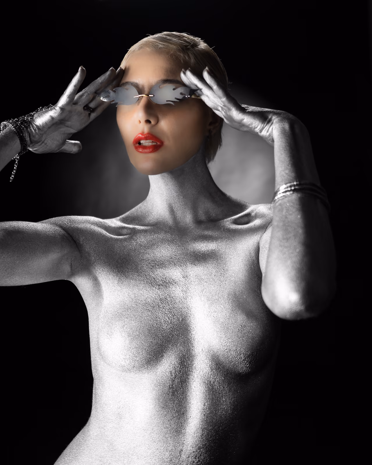 Una modella interamente dipinta d'argento tocca con le mani guantate gli occhiali da sole su sfondo nero.