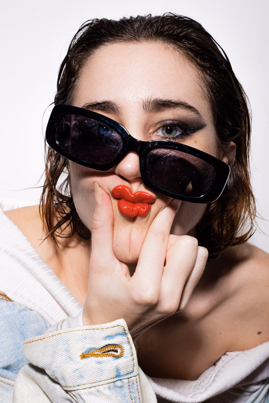 Primo piano di una donna con capelli bagnati e occhiali da sole neri abbassati, dito vicino alle labbra rosse atteggiate a bacio.