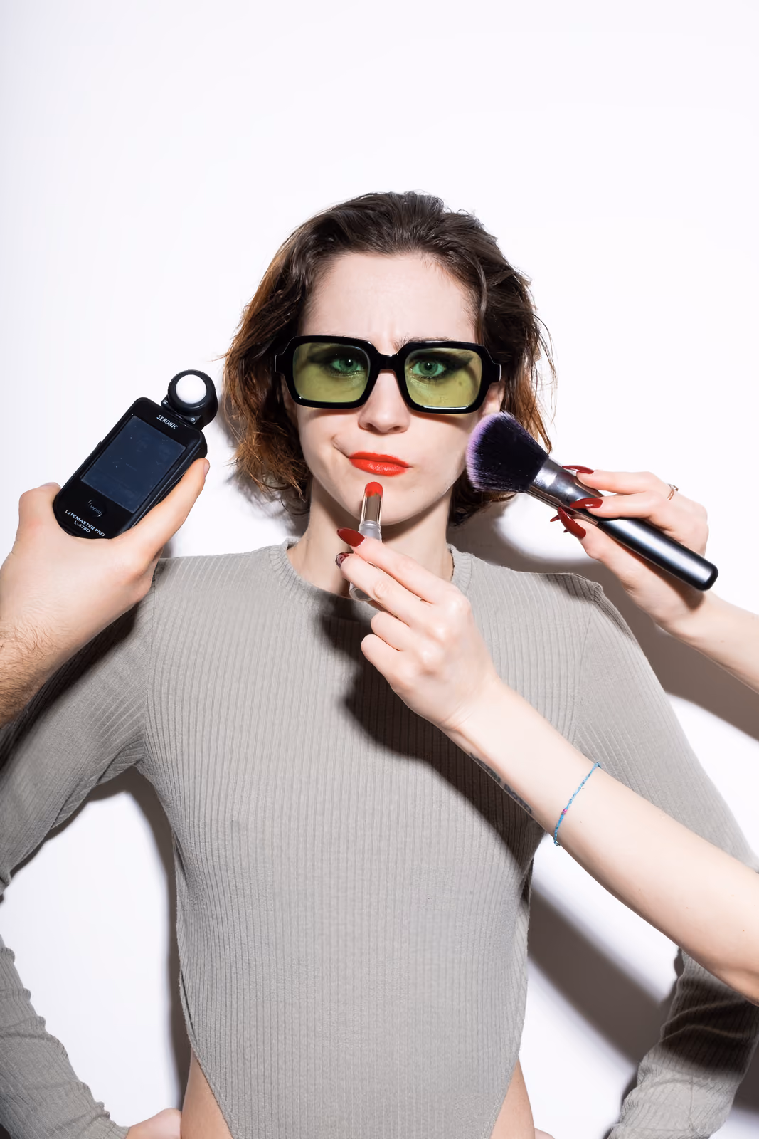 La modella con occhiali gialli tiene un rossetto rosso vicino alla bocca, mentre mani esterne reggono un misuratore di luce e un pennello per il trucco.