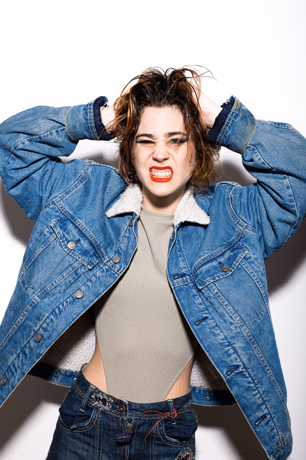 Una donna con le mani tra i capelli arruffati digrigna i denti, indossando una giacca di jeans oversize con collo in shearling.