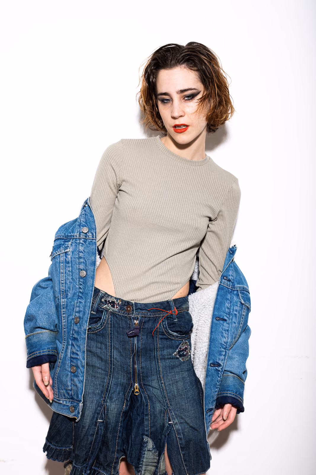 Una donna guarda di lato con la giacca di jeans che scivola dalle spalle, addome scoperto sopra una gonna di jeans plissettata.
