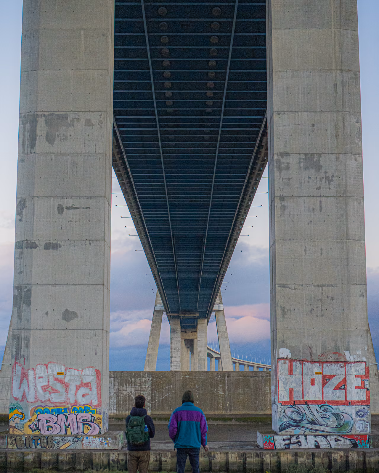 Due persone in piedi sotto gli enormi piloni in cemento di un ponte sospeso coperti di graffiti.