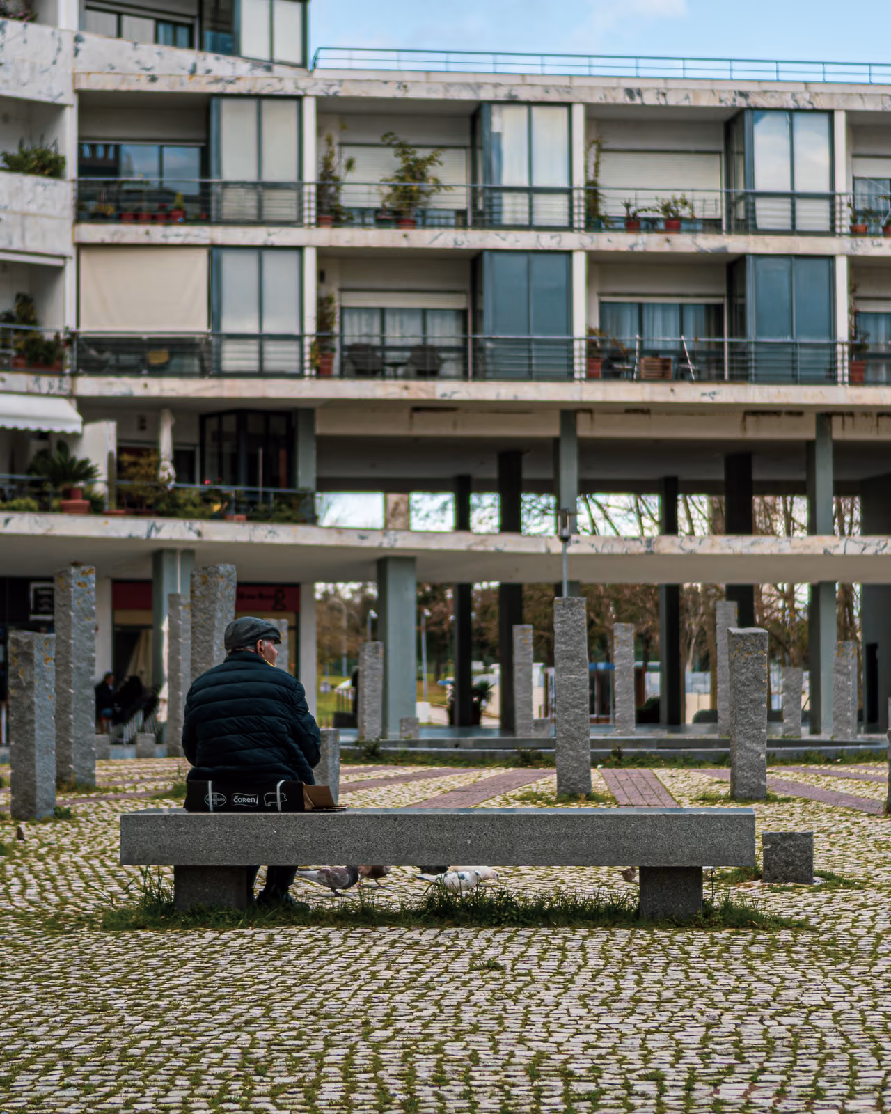 Un uomo anziano con berretto siede su una panchina di pietra in una piazza con piccioni, davanti a un edificio a terrazze.